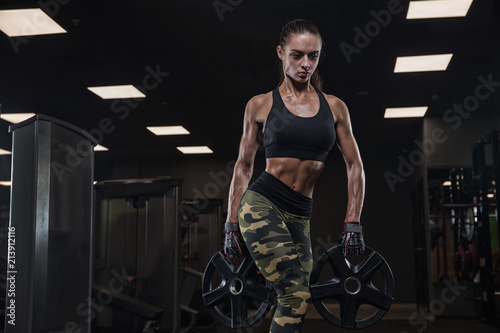 Young brunette woman, athlete bodybuilder doing exercises in a modern gym, against a dark background, in beautiful sportswear, camouflage. Concept-strength, beauty, training, diet, sports nutrition.