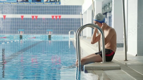 A young guy sits on the edge of the pool and puts a hat on his head.