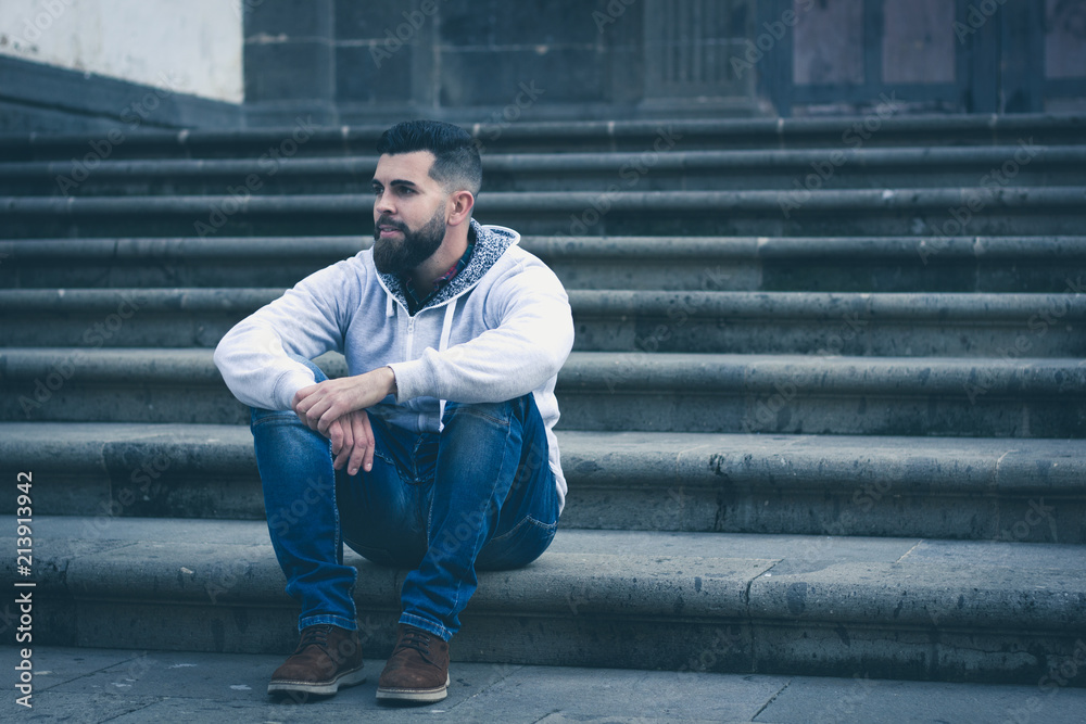 Thoughtful young man sitting outside church entrance stairs. Stylish male model with grey hoodie and denim jeans alone on the street. Cold blue tones in urban city Thoughtful young man sitting outside church entrance stairs. Stylish male model with grey hoodie and denim jeans alone on the street. Cold blue tones in urban city