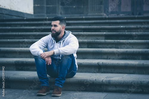 Thoughtful young man sitting outside church entrance stairs. Stylish male model with grey hoodie and denim jeans alone on the street. Cold blue tones in urban city