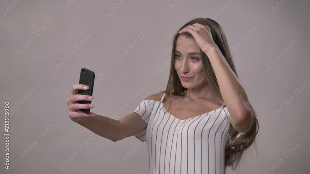 Young attractive shy girl is making selfie on smartphone, playing with hair, smiling, communication concept, grey background