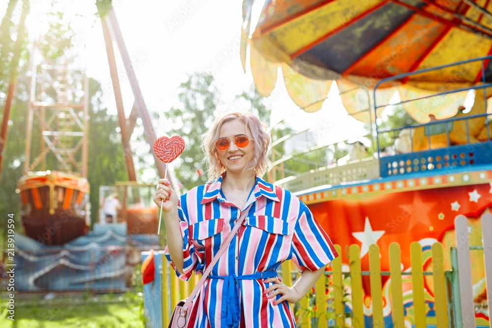 Bright summer image. The girl is blonde in  bright striped shirt dress walking in the amusement Park. Portrait of a woman on vacation. She holds a Lollipop in her hands and smiles cheerfully