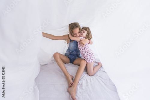Two beautiful sister kids playing under white sheets on bed. Fun indoors. Family love and lifestyle