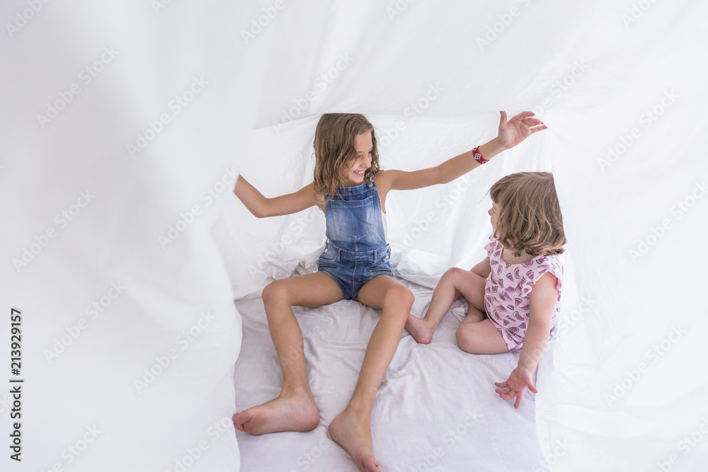 Two beautiful sister kids playing under white sheets on bed. Fun indoors. Family love and lifestyle