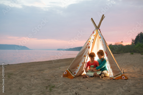 two children reading a book with flashlights in tent in the evening on the river bank. 