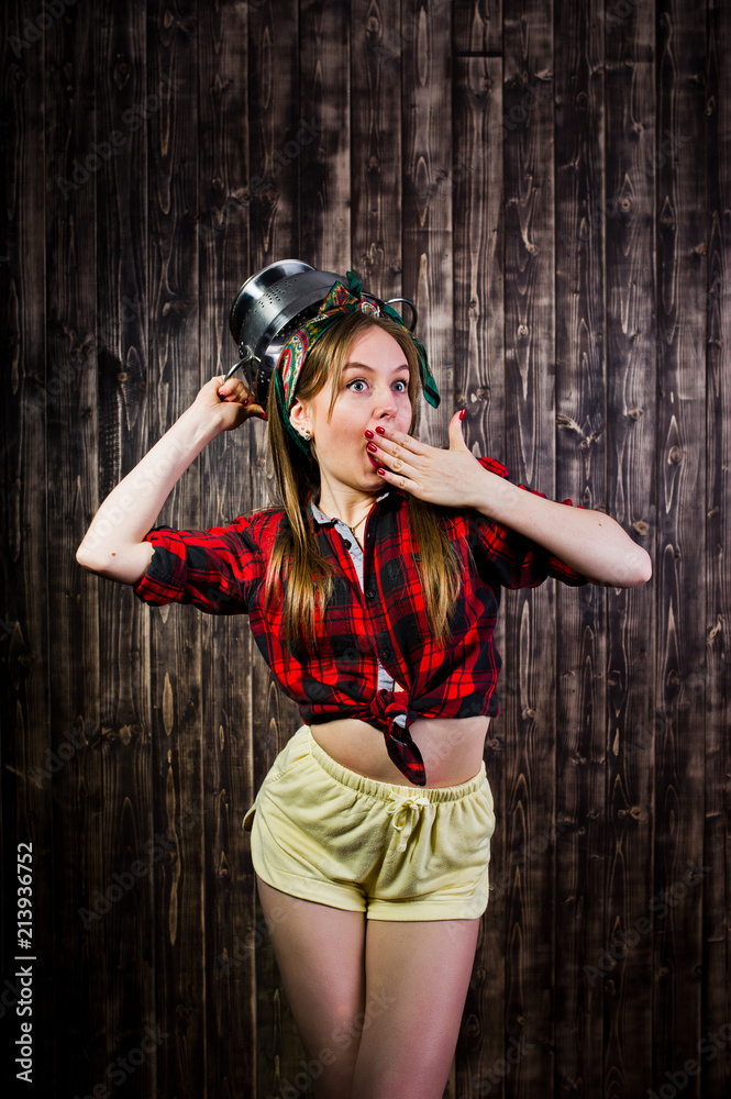 Obraz premium Young funny housewife in checkered shirt and yellow shorts pin up style with kitchen sieve on wooden background.