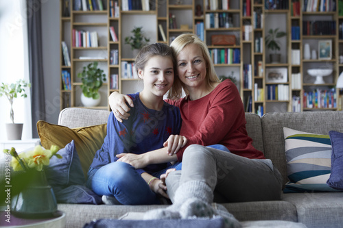 Mother and adolescent daughter sitting on couch with arms around