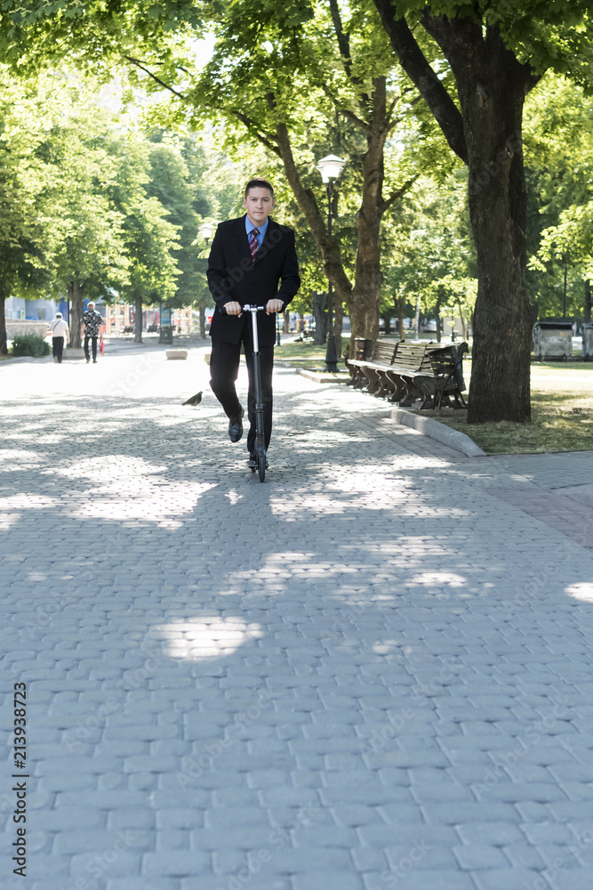 Businessman in suit is riding on a kick scooter in the city park