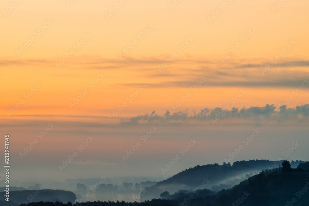 Morning mystic haze above broad valley of river. Gold glow from dawn in sky. Riverbank with forest under fog. Sunlight reflected in water at sunrise. Colorful atmospheric landscape of majestic nature.