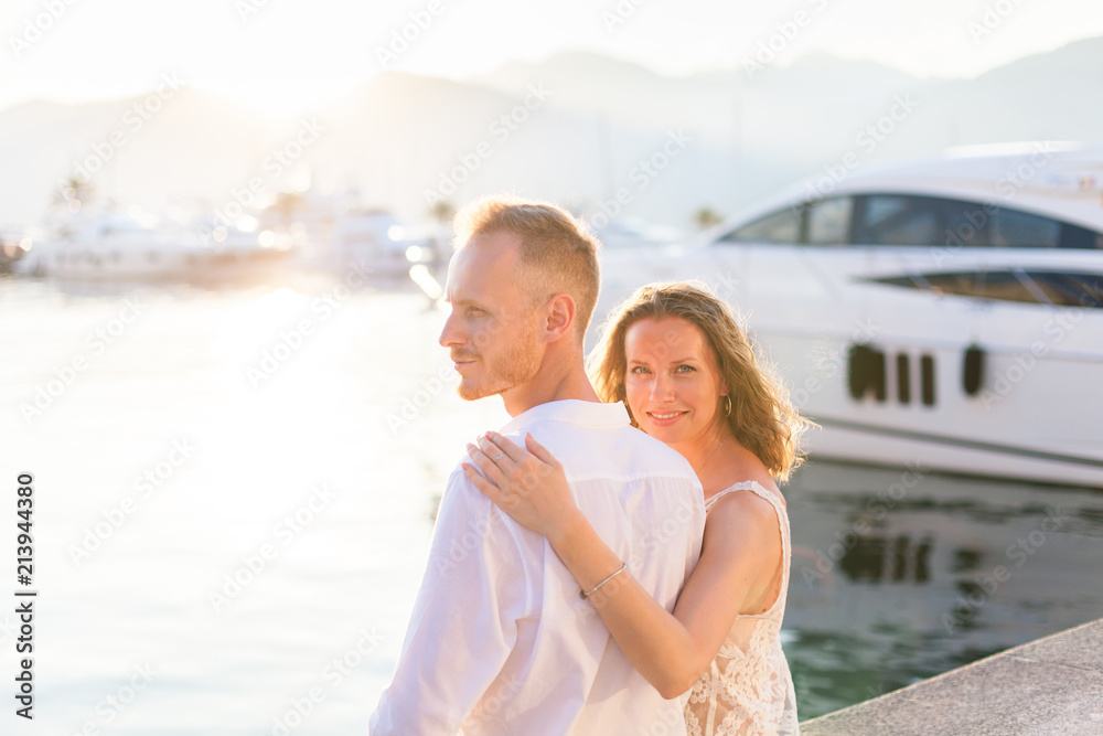 Couple of travelers are sitting in sea resort near yachts during romantic honeymoon vacation at sunset after wedding. Man and woman are happy tourists. Lovers are wearing in white clothes outdoor. Couple of travelers are sitting in sea resort near yachts during romantic honeymoon vacation at sunset after wedding. Man and woman are happy tourists. Lovers are wearing in white clothes outdoor.