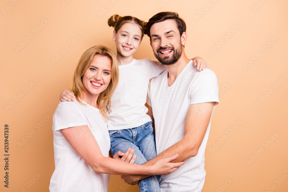 Portrait of cheerful adorable young family wearing white T-shirts. Lovely adorable mother and father holding their little daughter in their arms looking straight smiling on beige background