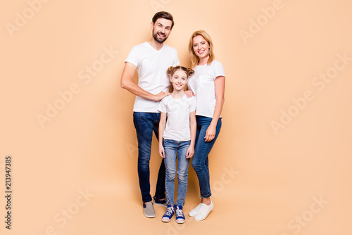 Full length portrait of adorable, attractive family standing together wearing white T-shirts and jeans on beige background. Bearded father and blonde mother keeping their hands on daughter's shoulders © deagreez