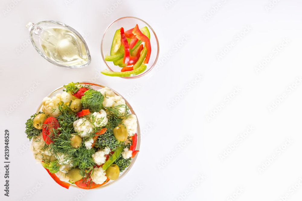 Fresh salad of cauliflower with tomato, broccoli, greens, olives and sweet pepper in a glass bowl along with a glass nipple on a white background. Top view. Copy space