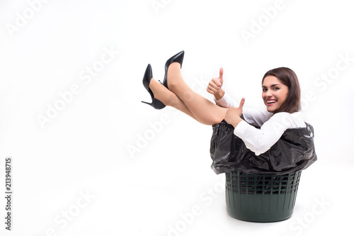 Happy woman sitting in trash bin showing thumbs up. Gorgeous dorky lady wearing white blouse and black heels sits in garbage can with her legs up. photo
