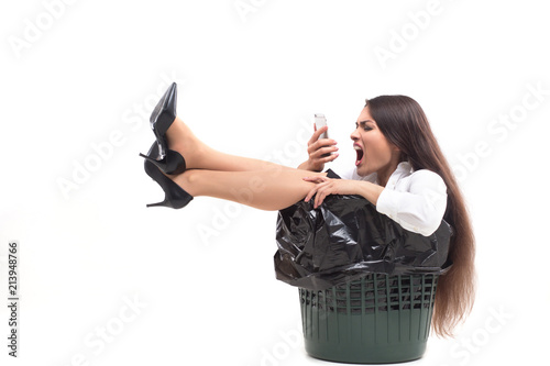 Lady screaming at her phone and sitting in trash bin. Crazy female shouts at her phone screen after having conversation while sitting in bucket meant for garbage. photo