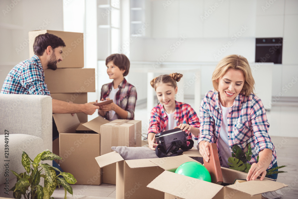 Young happy smiling family four persons unpacking carton boxes with stuff in light kitchen living room, moving to new flat Young happy smiling family four persons unpacking carton boxes with stuff in light kitchen living room, moving to new flat