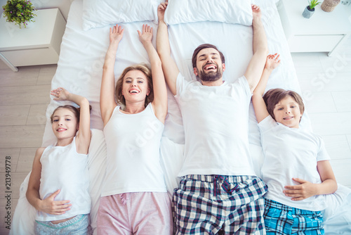 Young happy smiling family four persons wearing nightwear sleepwear lying together in bed hands up. Top above high angle view