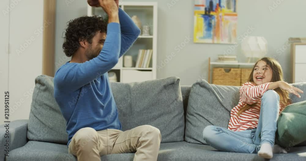 Caucasian father and small cute girl fighting with pillows on the couch in the living room. Indoors. Caucasian father and small cute girl fighting with pillows on the couch in the living room. Indoors.