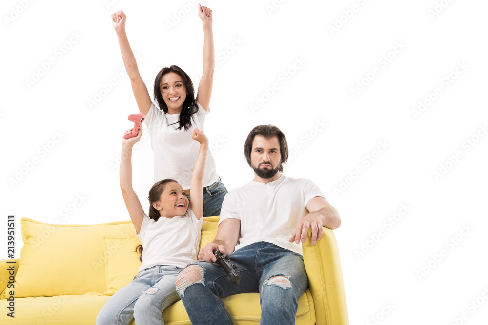 happy mother and daughter on yellow sofa playing video games with upset father near by isolated on white