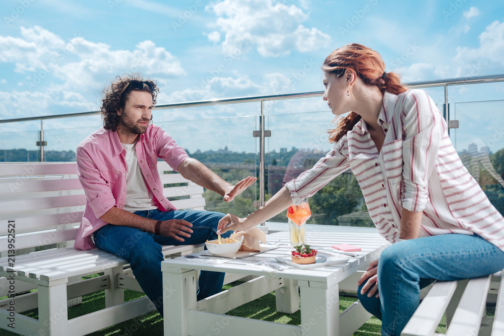 Obraz premium Lifting hand. Red-haired woman lifting her hand for trying fries of boyfriend while having lunch on summer terrace together