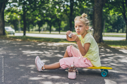 selective focus of adorable kid eating donut from dessert and looking away while sitting on skateboard at street