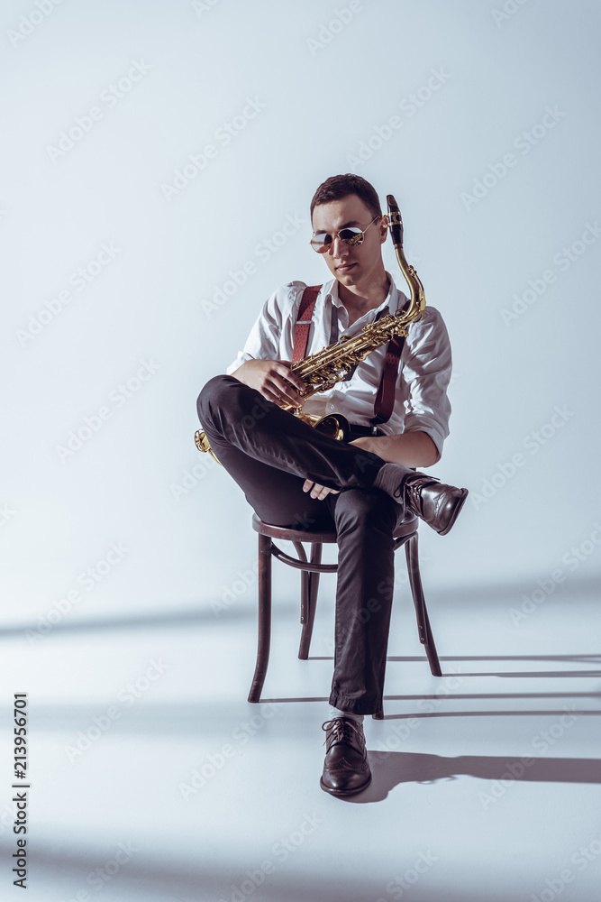 Fototapeta premium stylish young performer in sunglasses sitting on chair and holding saxophone on grey