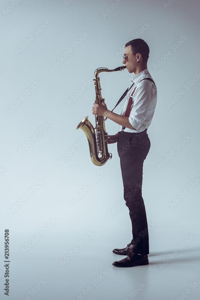Naklejka premium side view of stylish young professional musician playing saxophone on grey