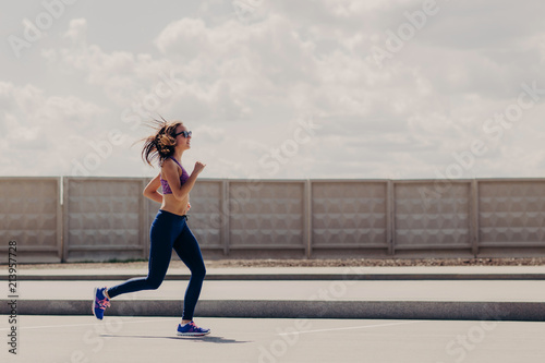 Horizontal shot sportive woman joggs in open air, enjoys sport activities, wears sport clothes, concentrated into distance, has running workout. Female runner motivates to go in sport Stock Photo