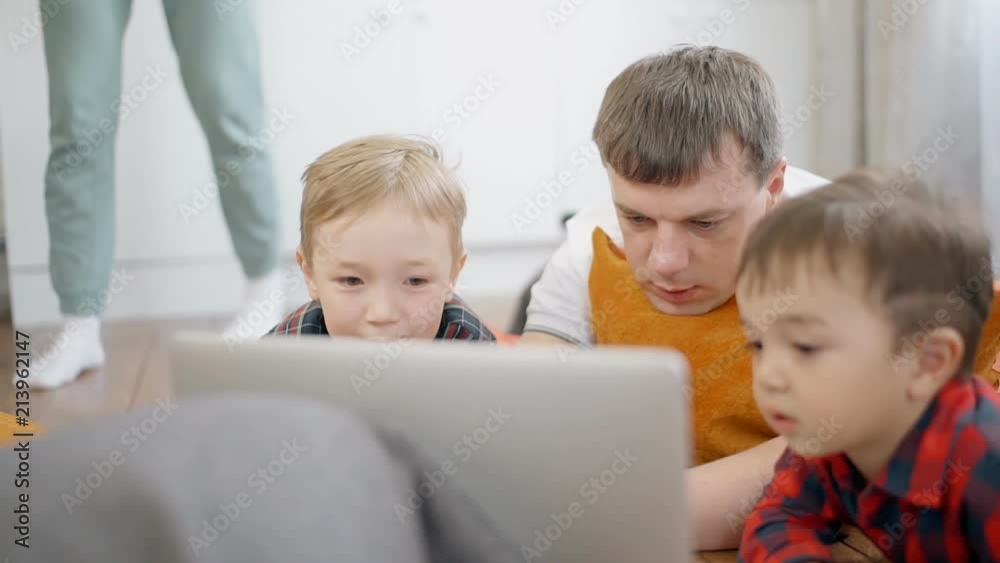 Young man with two children using laptop, lying on floor in home interior, caring father and little sons are looking at monitor, talking with smiles in bright apartment. Concept: loving family Young man with two children using laptop, lying on floor in home interior, caring father and little sons are looking at monitor, talking with smiles in bright apartment. Concept: loving family