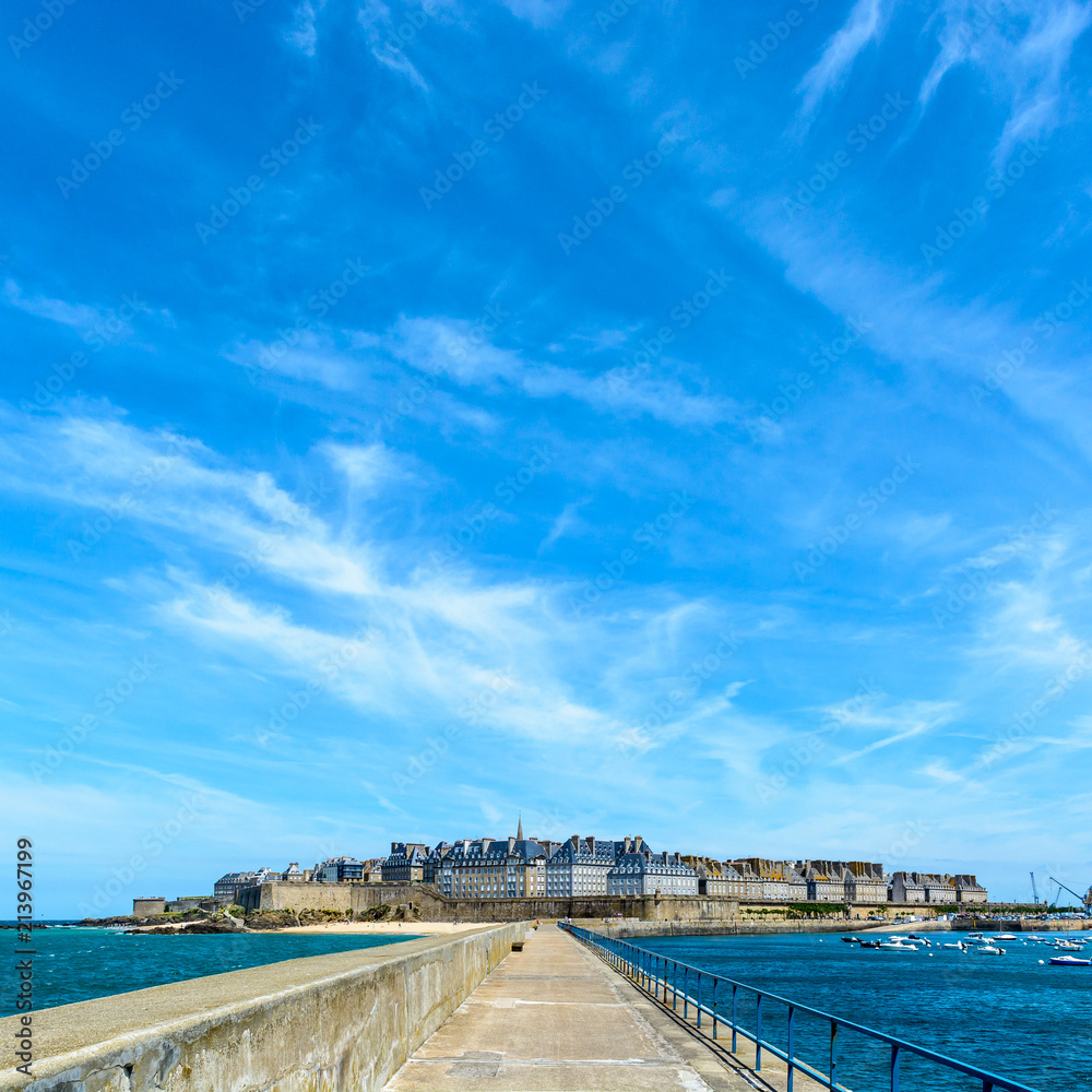 General view of the walled city of Saint-Malo in Brittany, France, with the granite residential buildings sticking out above the rampart, seen from the breakwater under a summer blue sky.
