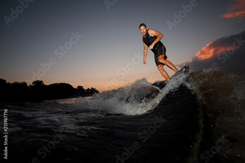 Athletic wakesurfer jumping on board riding down the river waves at the sunset