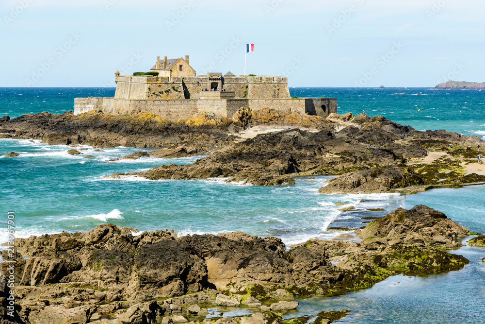 The Fort National built by french military architect Vauban on a tidal island, seen from the city of Saint-Malo, France, with the french flag blowing in the wind and rocks in the sea in the foreground The Fort National built by french military architect Vauban on a tidal island, seen from the city of Saint-Malo, France, with the french flag blowing in the wind and rocks in the sea in the foreground