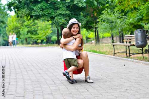 A young beautiful mother in a red skirt and white T-shirt hugs her two-year-old son in the park.
