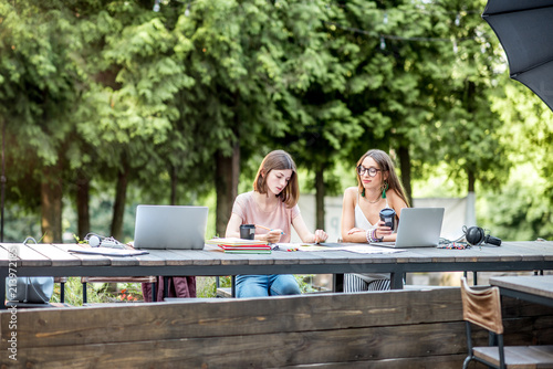 Young girlfriends sitting together studying with laptops and coffee at the outdoor park cafe