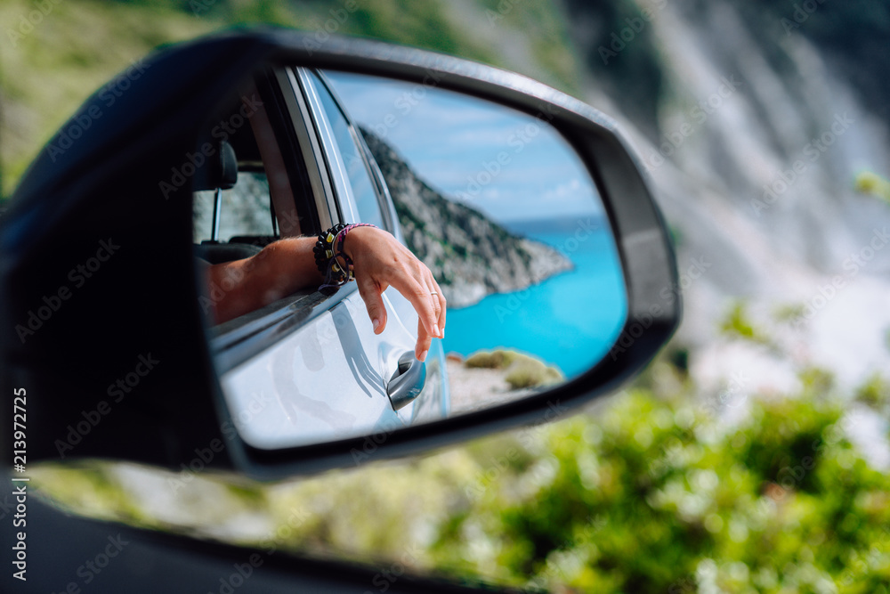 Brown female hand in the car side view mirror. Blue mediterranean sea and white rocks in background