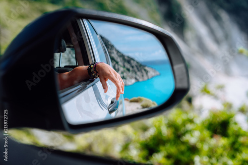 Brown female hand in the car side view mirror. Blue mediterranean sea and white rocks in background © Igor Tichonow