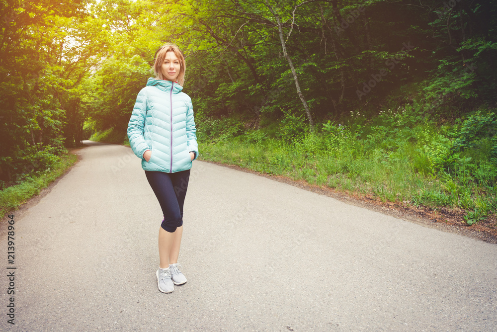 Portrait of an attractive sports girl blonde in a light running down jacket dressing bluetooth headphones with music or the sounds of nature while on a forest road