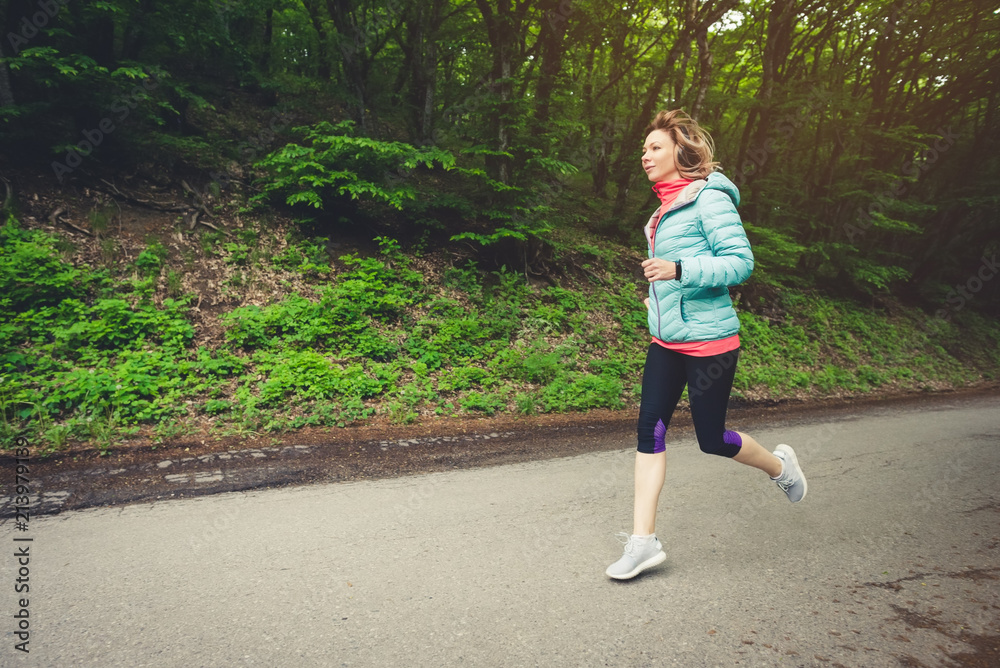 Young fitness blonde woman in headphones running at morning caucasian forest trail in sun light Young fitness blonde woman in headphones running at morning caucasian forest trail in sun light