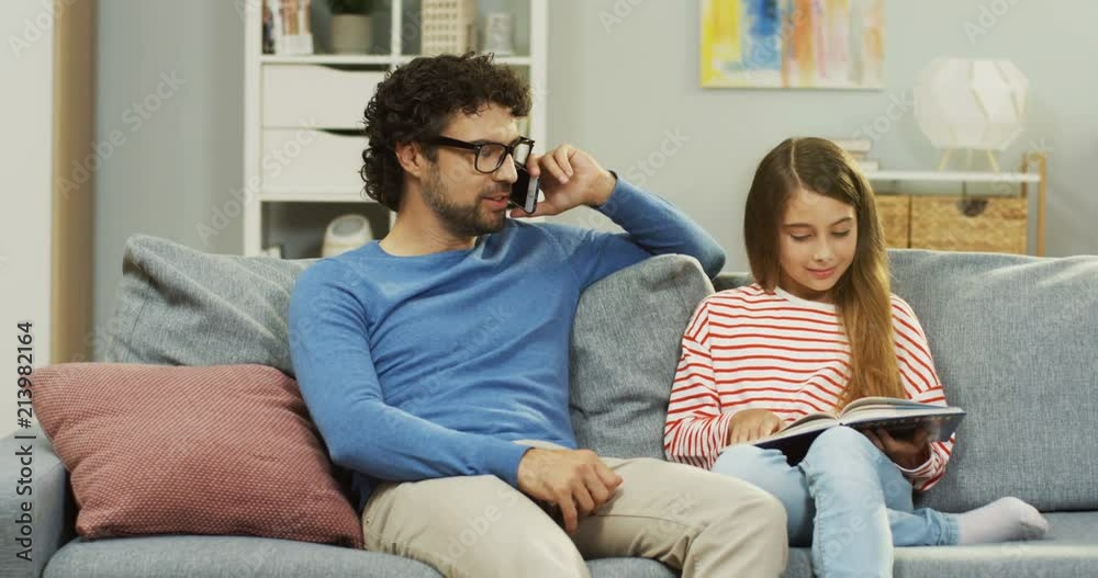 Little schoolgirl reading a textbook on the couch while her daddy talking on the phone next to her. Indoors.