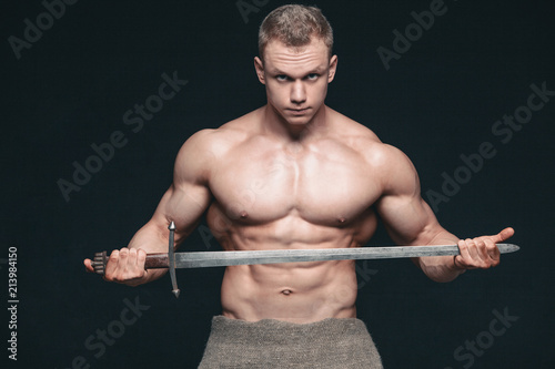 Bodybuilder man posing with a sword isolated on black background. Serious shirtless man demonstrating his mascular body © satyrenko