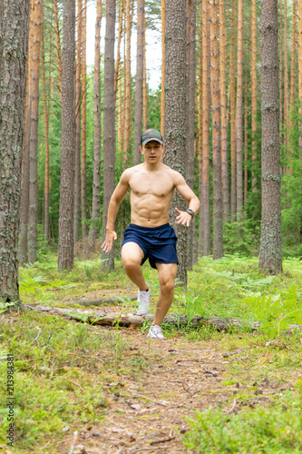 A young man with a muscular bare chest  in shorts and a cap  runs along a pine forest against the background of tree trunks