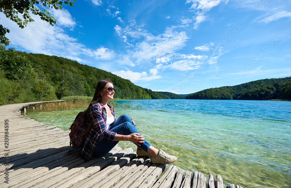 Young smiling tourist woman in sunglasses and with backpack sitting on wooden bridge, enjoying beautiful view, colorful spring panorama of lit by sun green forest and blue lake under clear sky