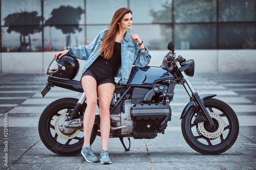 Portrait of a young beautiful brunette girl dressed in a shorts and jeans jacket sitting on a custom-made retro motorcycle in city, looking away.