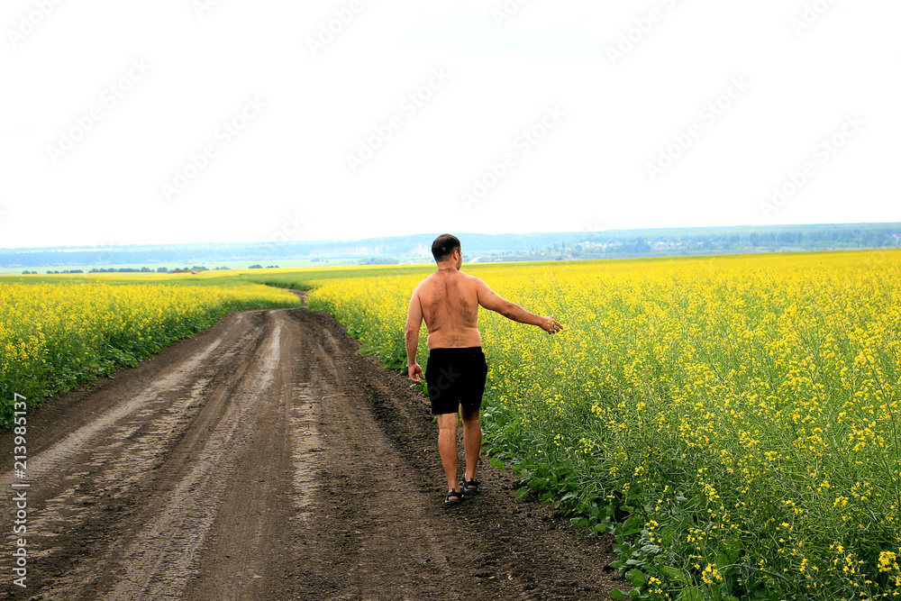 A young man walks along a dirt road, along a field of yellow flowers.