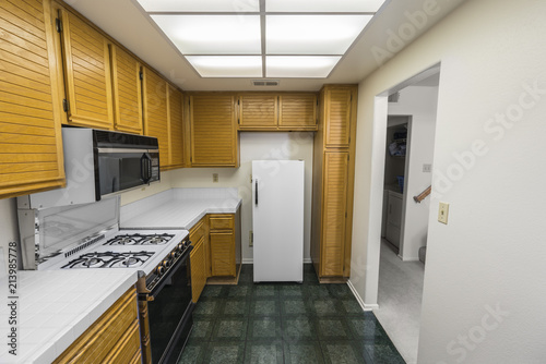 Old 1980s condo kitchen with oak cabinets, tile countertops, gas stove and green flooring. 