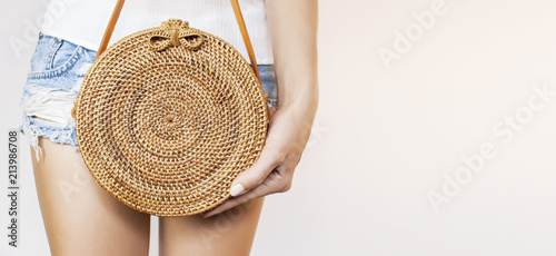 Young girl in denim shorts and a white sweatshirt with a trendy stylish rattan bag against the backdrop of a light wall. Rattan handbag, ecobags from Bali. Eco-bag concept, trendy bamboo bag.  photo