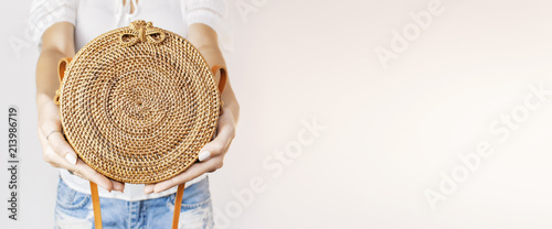 Young girl in denim shorts and a white sweatshirt with a trendy stylish rattan bag against the backdrop of a light wall. Rattan handbag, ecobags from Bali. Eco-bag concept, trendy bamboo bag.  photo