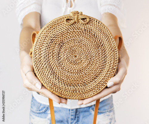 Young girl in denim shorts and a white jacket opens a stylish natural rattan bag against the backdrop of a light wall. Rattan handbag, ecobags from Bali. Eco-bag concept, trendy bamboo bag. photo