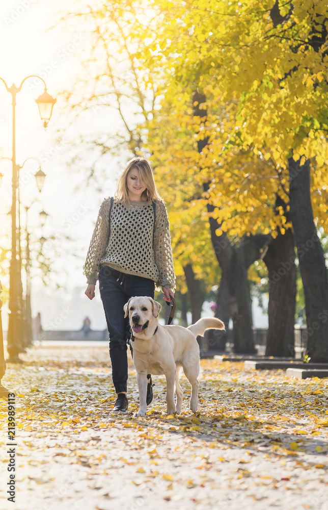 A blonde is walking with a labrador in the park.