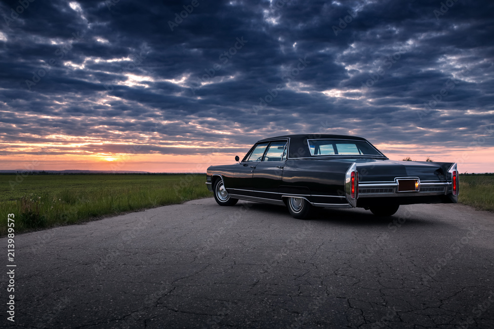 Fototapeta premium Czarny retro vintage muscle car jest zaparkowany na wiejskiej drodze asfaltowej na złoty zachód słońca